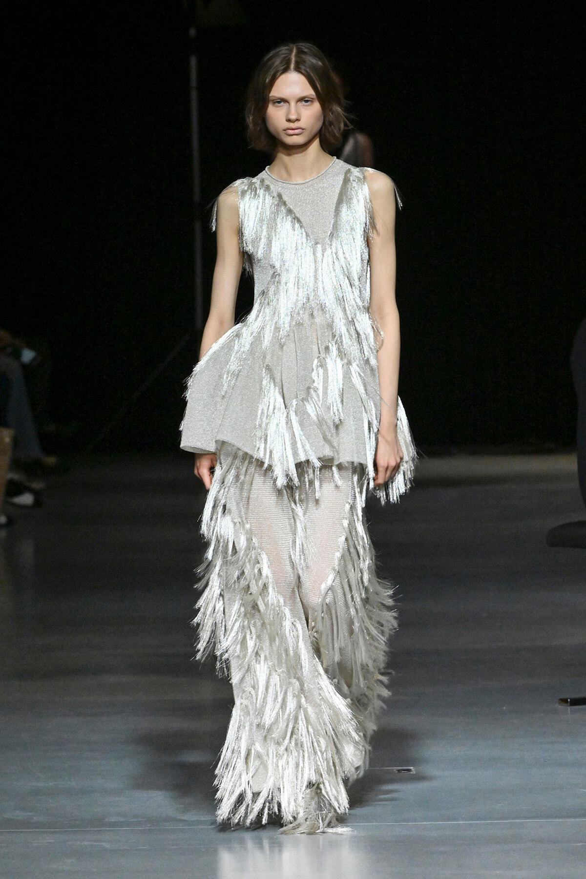 Model walks the runway in a silver fringe gown with cascading tassels on a dark catwalk