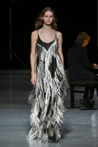 Model walking a fashion runway in a black slip dress adorned with long silver fringe tassels cascading from the neckline to the hem. A pianist sits in the background.