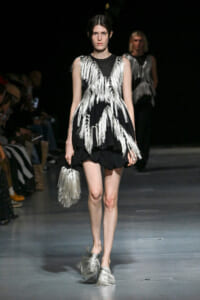 Model on a runway wearing a black sleeveless dress with long silver fringe and a matching fringed silver handbag.