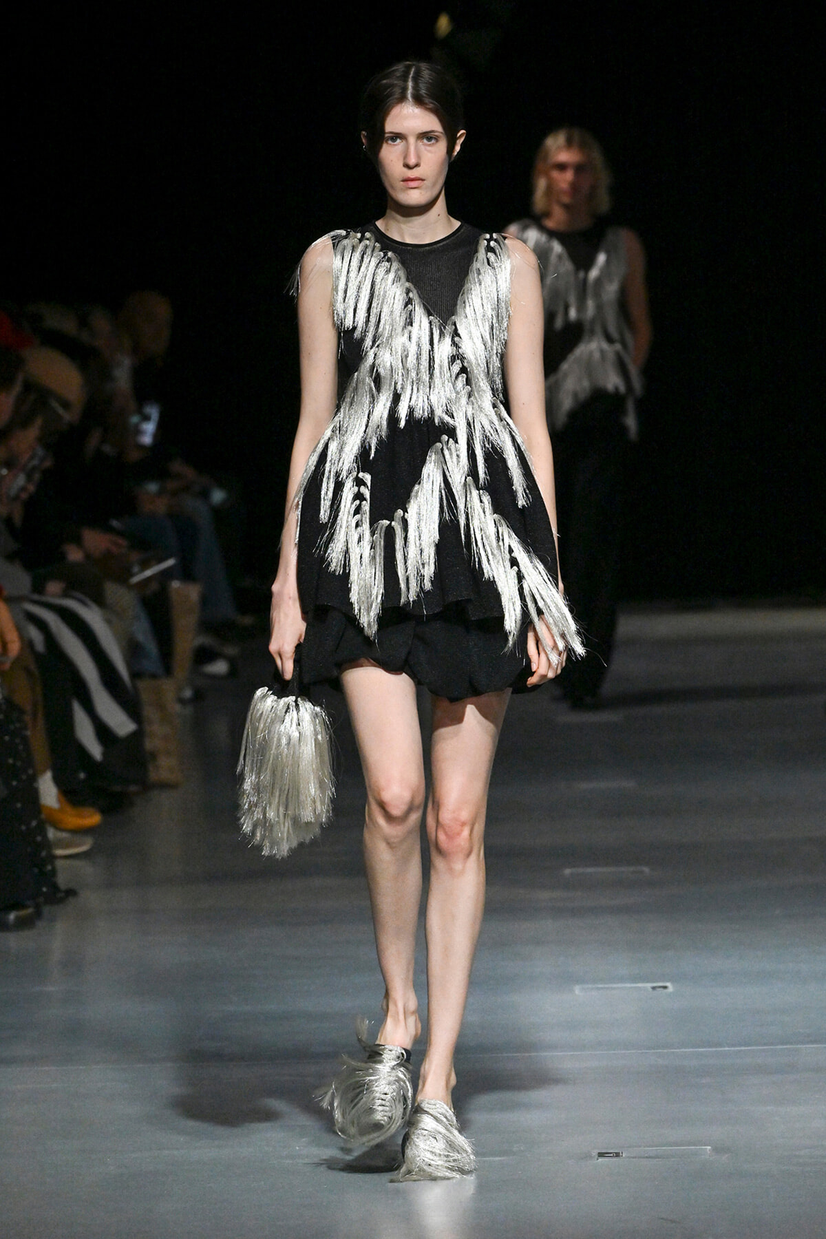 Model on a runway wearing a black sleeveless dress with long silver fringe and a matching fringed silver handbag.