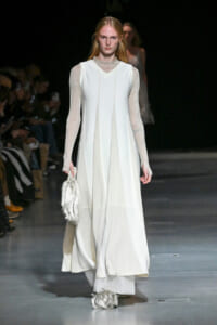 Model walks the runway in a white, floor-length gown with sheer, glittery sleeves, carrying a silver fringed bag at her side