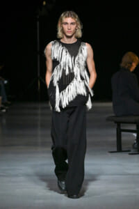 Male model walks the runway in a black sleeveless top with bold white fringe tassels over loose black pants.