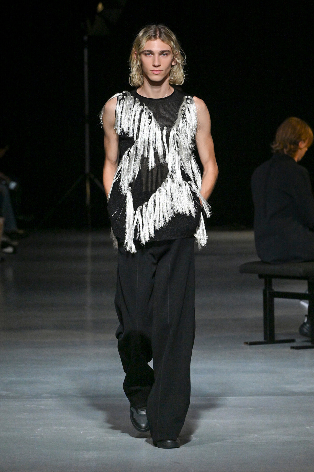 Male model walks the runway in a black sleeveless top with bold white fringe tassels over loose black pants.