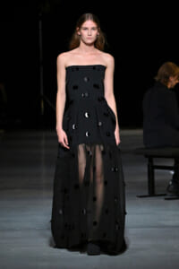 Model walking a runway in a strapless black gown adorned with round black sequins and a sheer, floor-length overlay color-darkened hem.