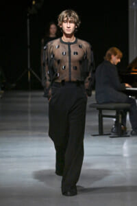 Male model walks the runway in a sheer black blouse with circular appliqués and high-waisted black trousers, pianist visible in the background.