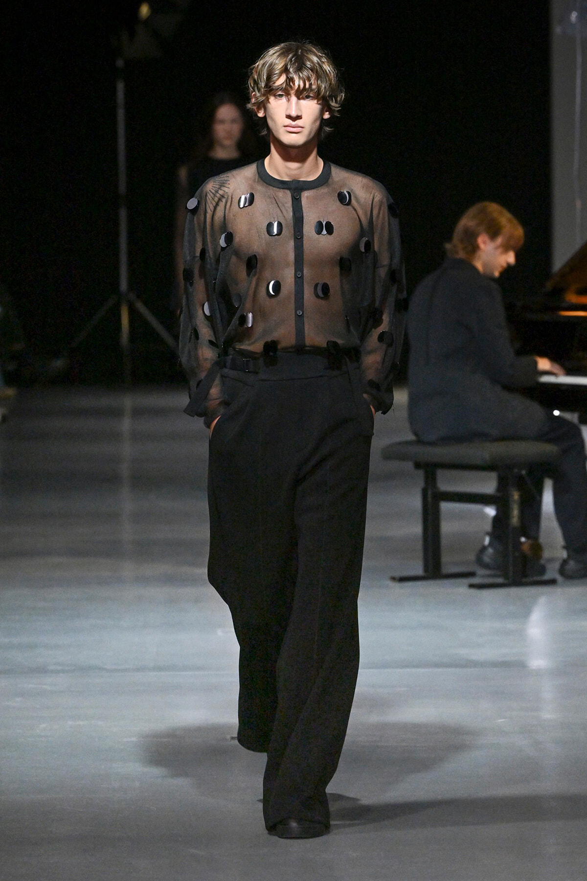 Male model walks the runway in a sheer black blouse with circular appliqués and high-waisted black trousers, pianist visible in the background.