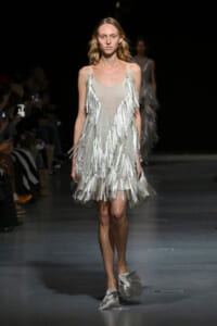 Model walking the runway in a shimmering silver fringe dress with long fringe strands cascading from the shoulders and hem.