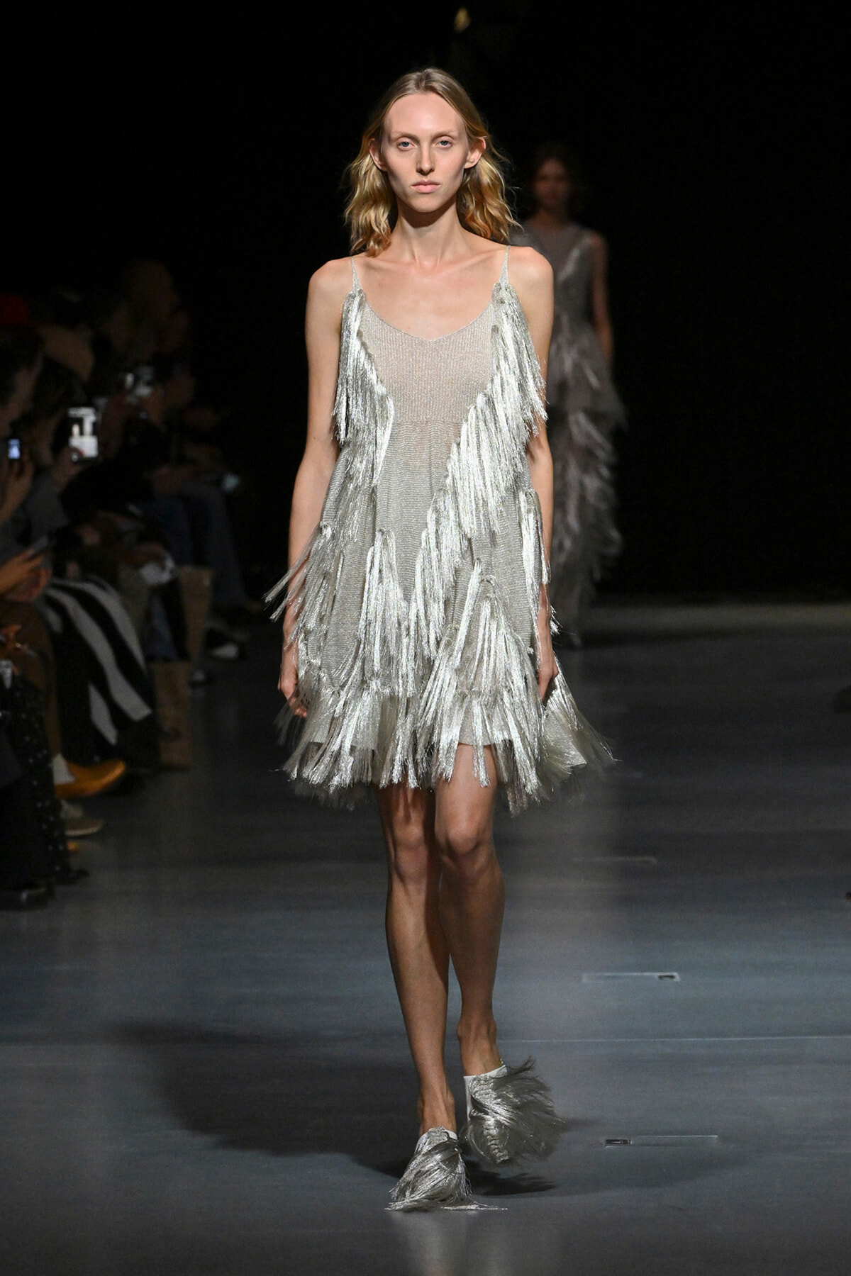 Model walking the runway in a shimmering silver fringe dress with long fringe strands cascading from the shoulders and hem.
