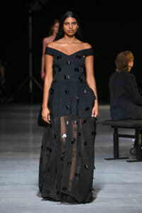 Model walking a runway in an off-the-shoulder black gown with a sheer skirt and scattered circular sequins/embellishments.