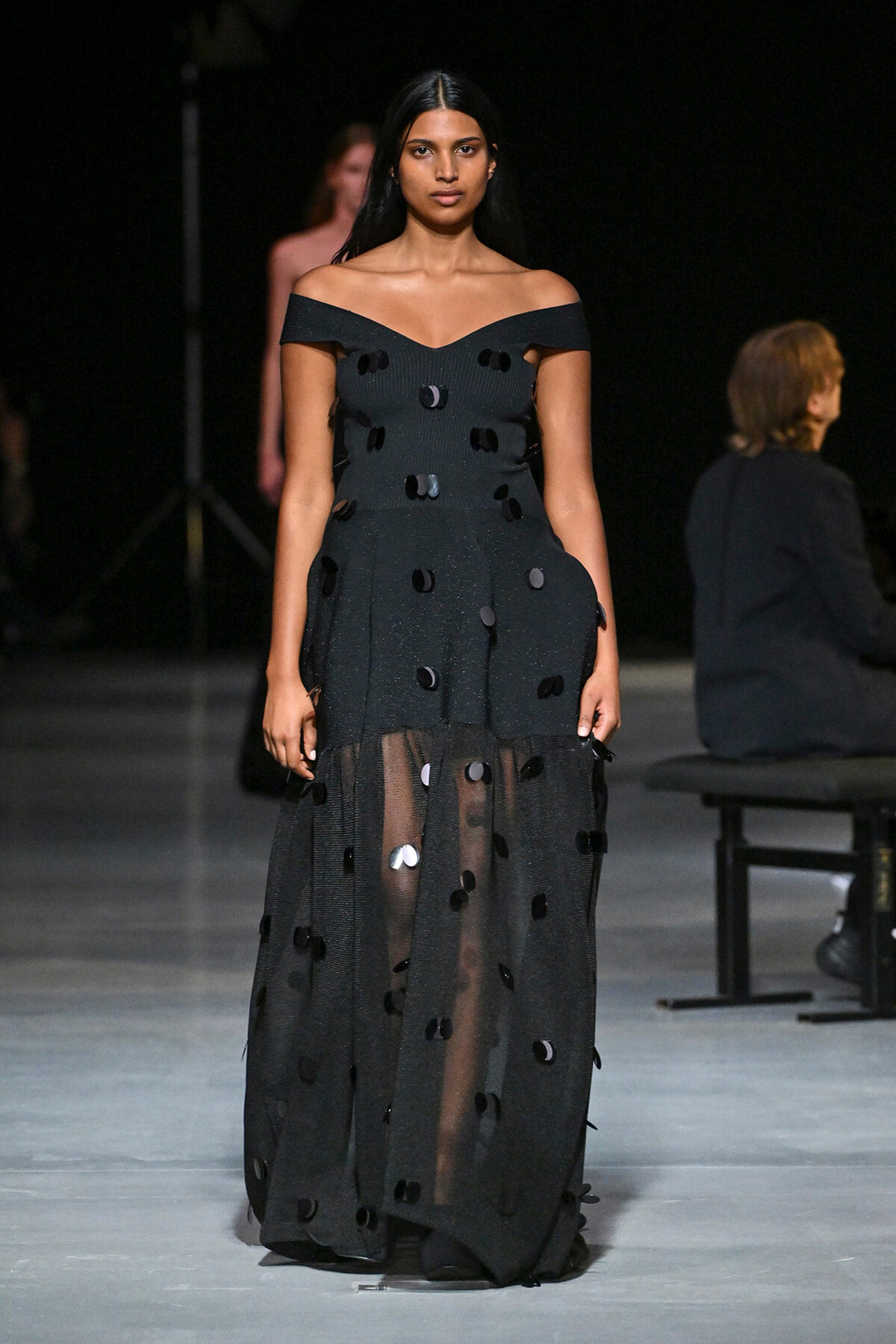 Model walking a runway in an off-the-shoulder black gown with a sheer skirt and scattered circular sequins/embellishments.