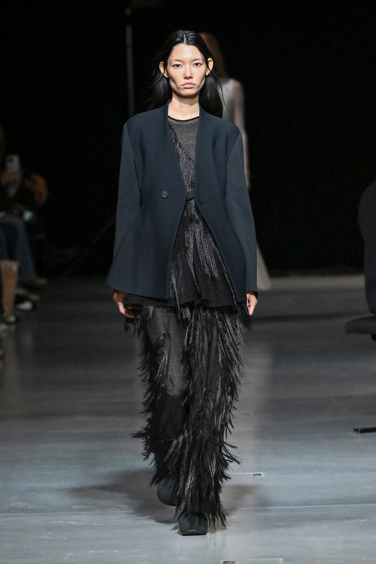 Model walks a fashion runway in a dark navy blazer over a sheer black fringe dress; audience blurred in the background.