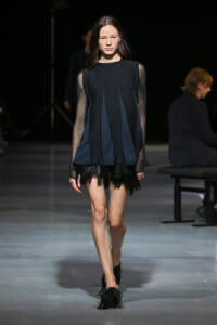 Model walking a runway in a navy blue, sleeved dress with sheer mesh sleeves and a fringed black hem.