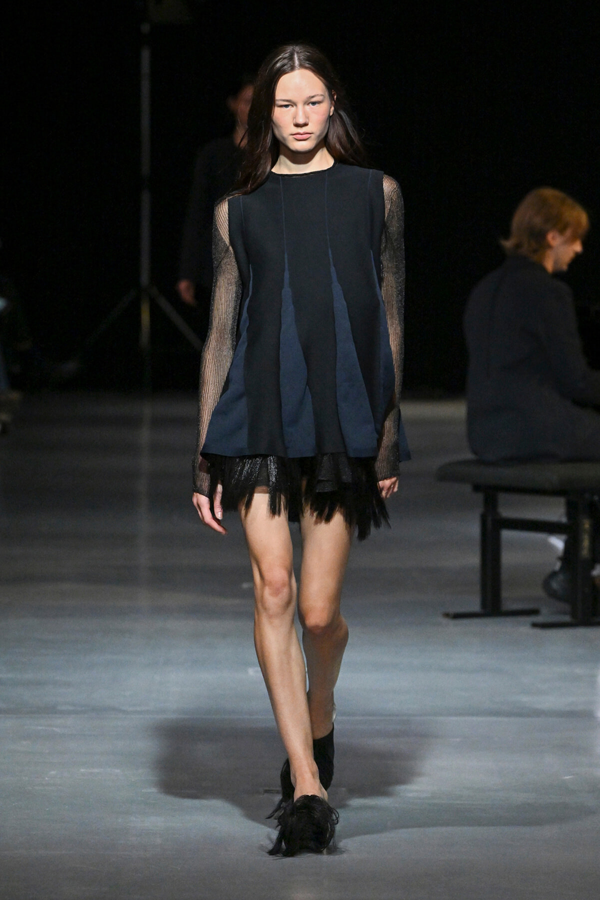 Model walking a runway in a navy blue, sleeved dress with sheer mesh sleeves and a fringed black hem.