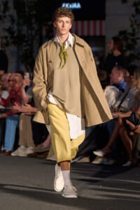 Male model walking a runway in a beige trench over a layered tan sweater and shirt, with pale yellow shorts and white sneakers, lime scarf at the neckline.