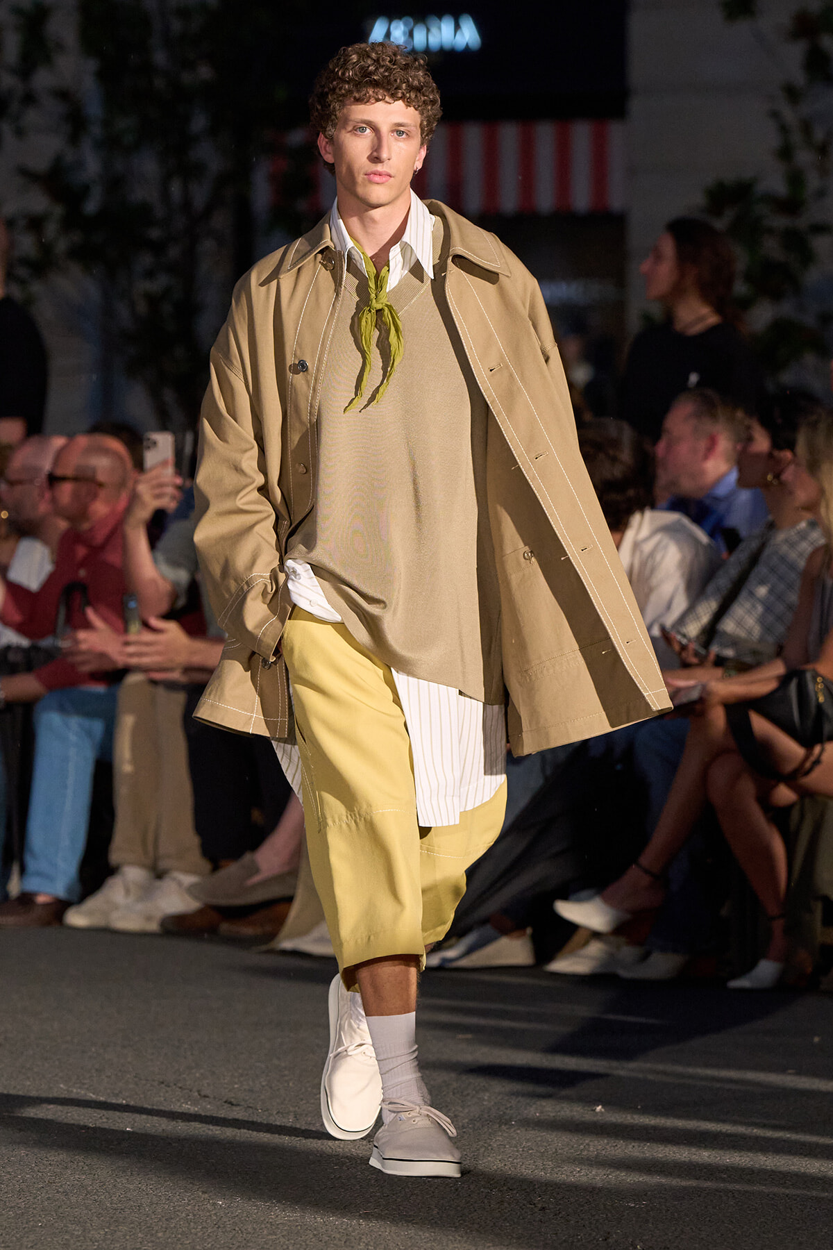 Male model walking a runway in a beige trench over a layered tan sweater and shirt, with pale yellow shorts and white sneakers, lime scarf at the neckline.