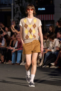 Runway model wearing a beige argyle sweater vest over a white T-shirt, brown suede shorts, white socks, navy sneakers, and red-orange sunglasses, carrying a small bag as the audience watches.