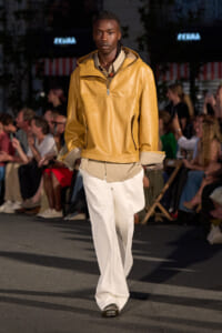 Male model walking a runway in a mustard leather jacket, beige layered shirt, and wide white pants, with an audience in the background.