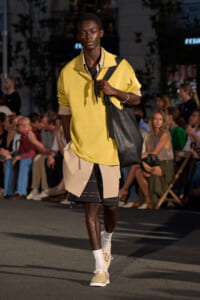 Male model walking a street-style runway in a loose yellow polo over beige shirt, layered shorts, and a large black shoulder bag.