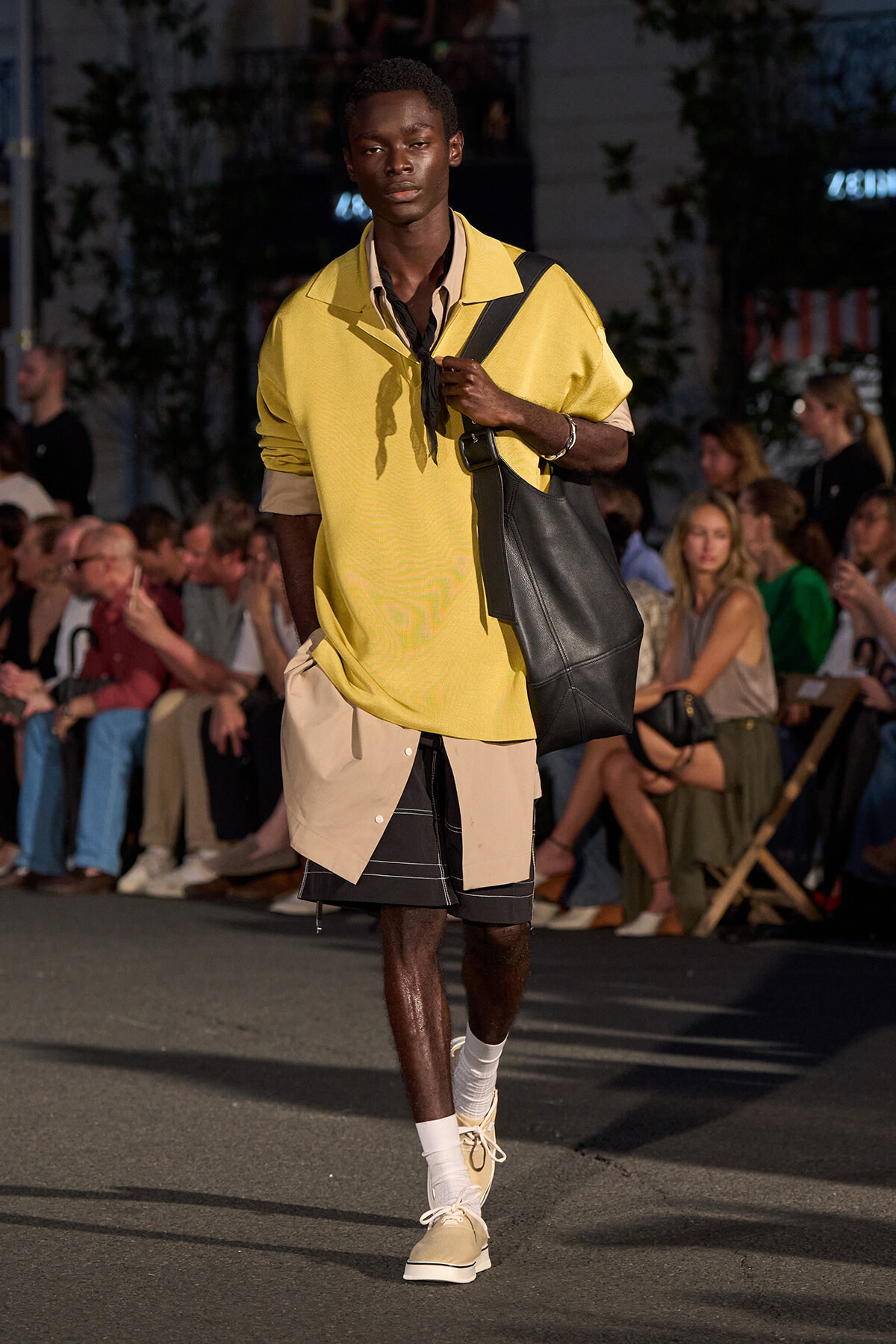 Male model walking a street-style runway in a loose yellow polo over beige shirt, layered shorts, and a large black shoulder bag.
