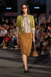 Model walking a fashion runway in a lime-green jacket over a striped blouse and tan suede pencil skirt with a large belt, carrying a small tan bag.