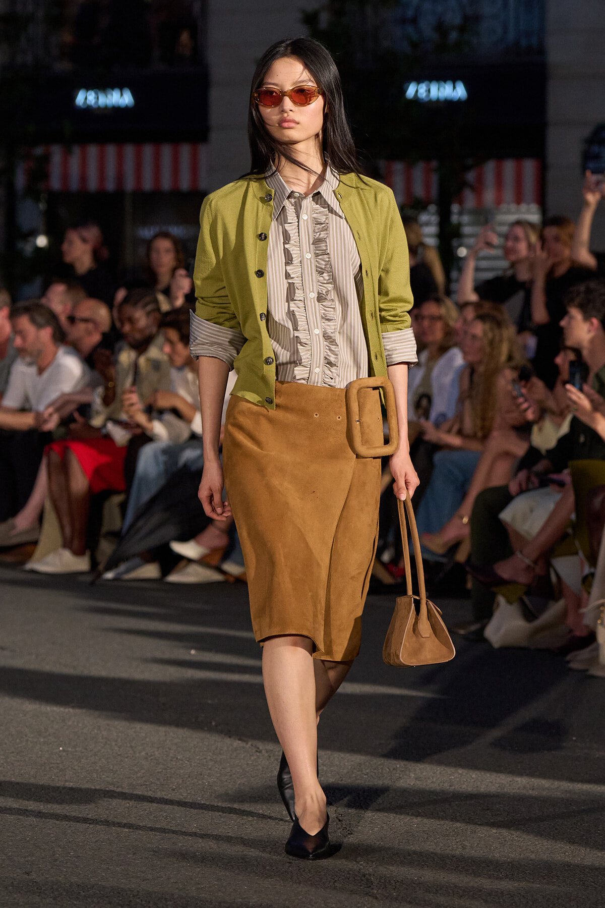 Model walking a fashion runway in a lime-green jacket over a striped blouse and tan suede pencil skirt with a large belt, carrying a small tan bag.