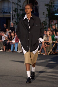 Model walks a street-style runway wearing an oversized black utility jacket over a white shirt, paired with beige shorts, white socks, and black shoes as crowds watch a fashion show.