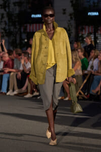 Model walks a runway in a mustard-yellow suede jacket over a yellow top and gray knee-length skirt with a front slit, beige shoes, and an olive handbag.