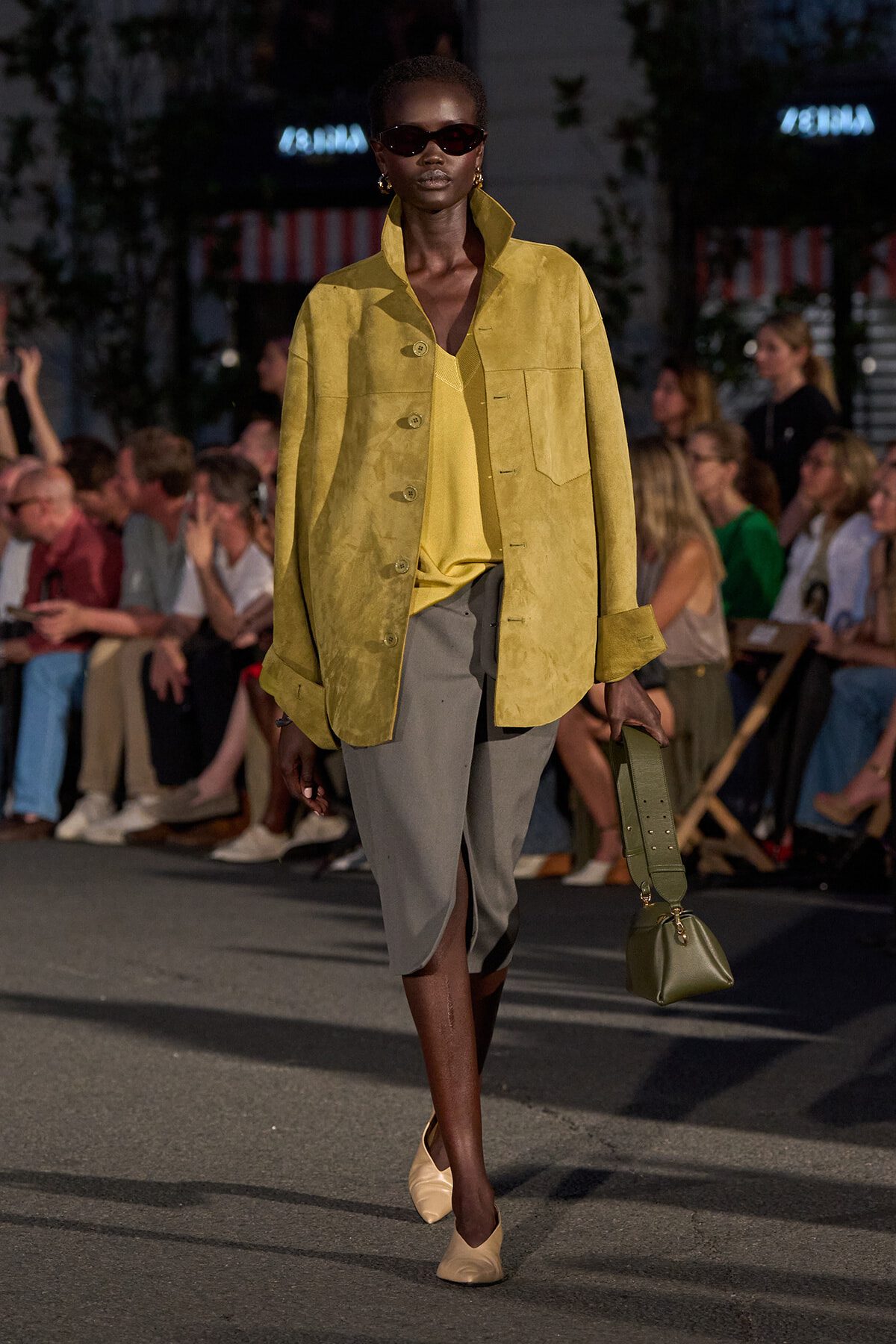 Model walks a runway in a mustard-yellow suede jacket over a yellow top and gray knee-length skirt with a front slit, beige shoes, and an olive handbag.