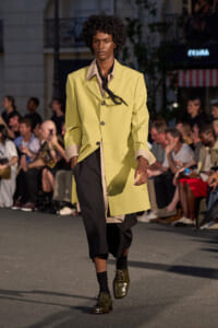 Male model on a runway wearing a pale yellow coat over black pants with a black scarf, walking past an audience.