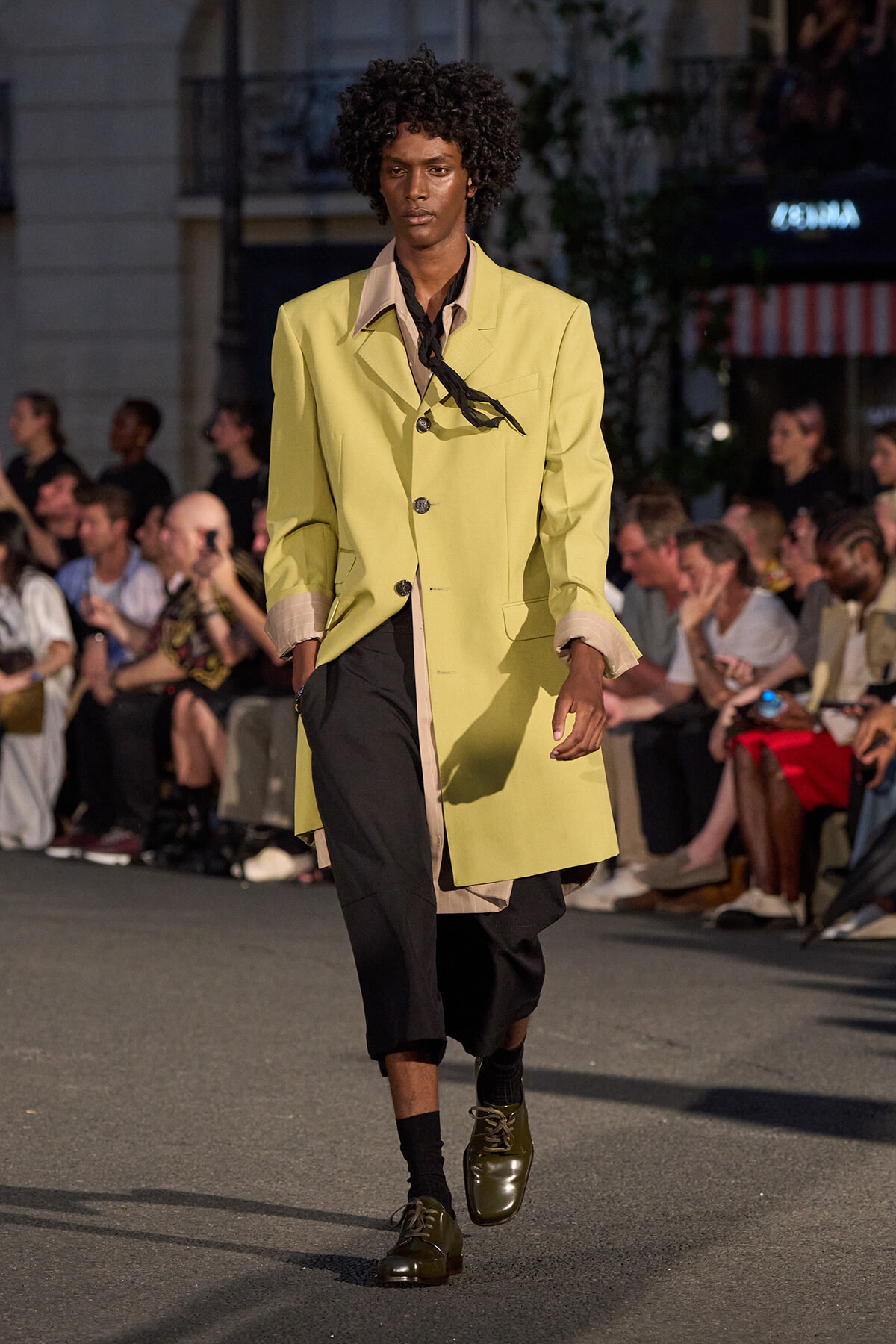 Male model on a runway wearing a pale yellow coat over black pants with a black scarf, walking past an audience.