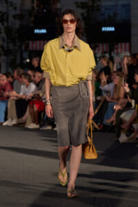 Model walking a runway in a yellow oversized button-down shirt, gray knee-length skirt with front slit, green sandals, red sunglasses, and a tan handbag.