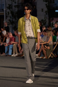 Male model on a runway wearing a lime-green jacket over a striped shirt, gray wide-leg trousers, white sneakers, and dark sunglasses.