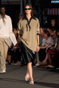 Model walks a fashion runway in a beige tunic with a black neck scarf, a black leather skirt, white sandals, and dark sunglasses.
