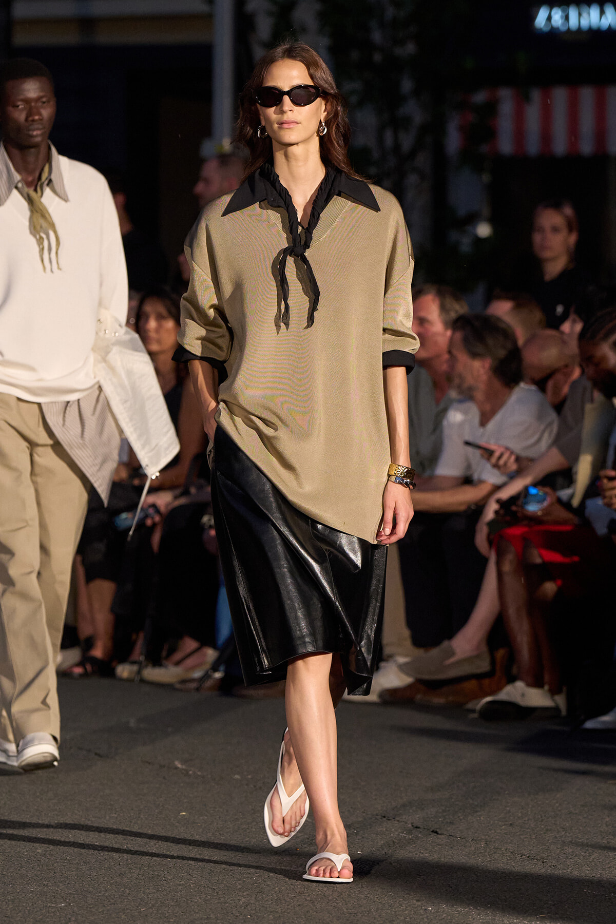 Model walks a fashion runway in a beige tunic with a black neck scarf, a black leather skirt, white sandals, and dark sunglasses.