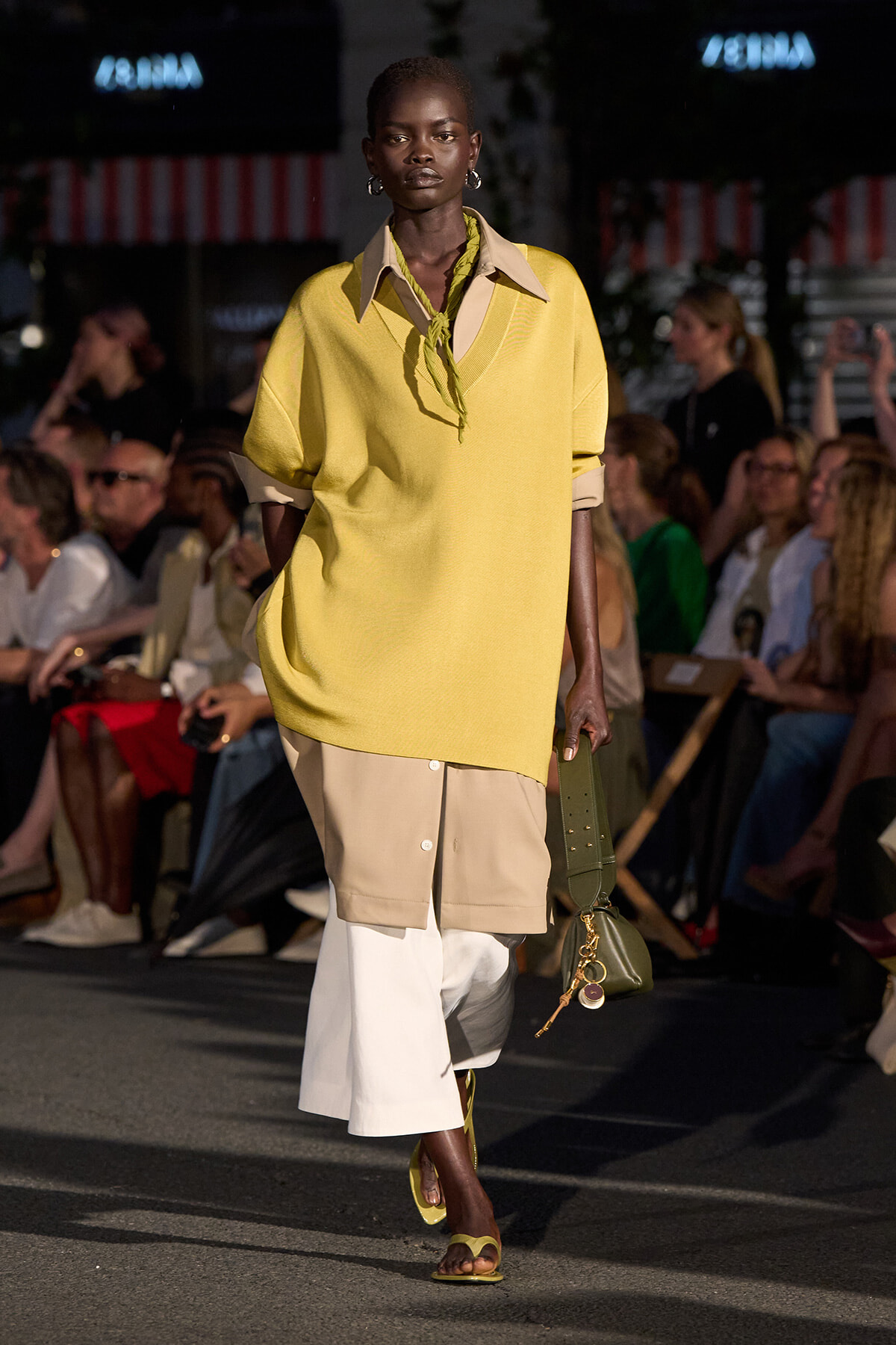 Runway model in an oversized yellow sweater layered over a beige shirt-dress with white cropped pants, carrying a green handbag.