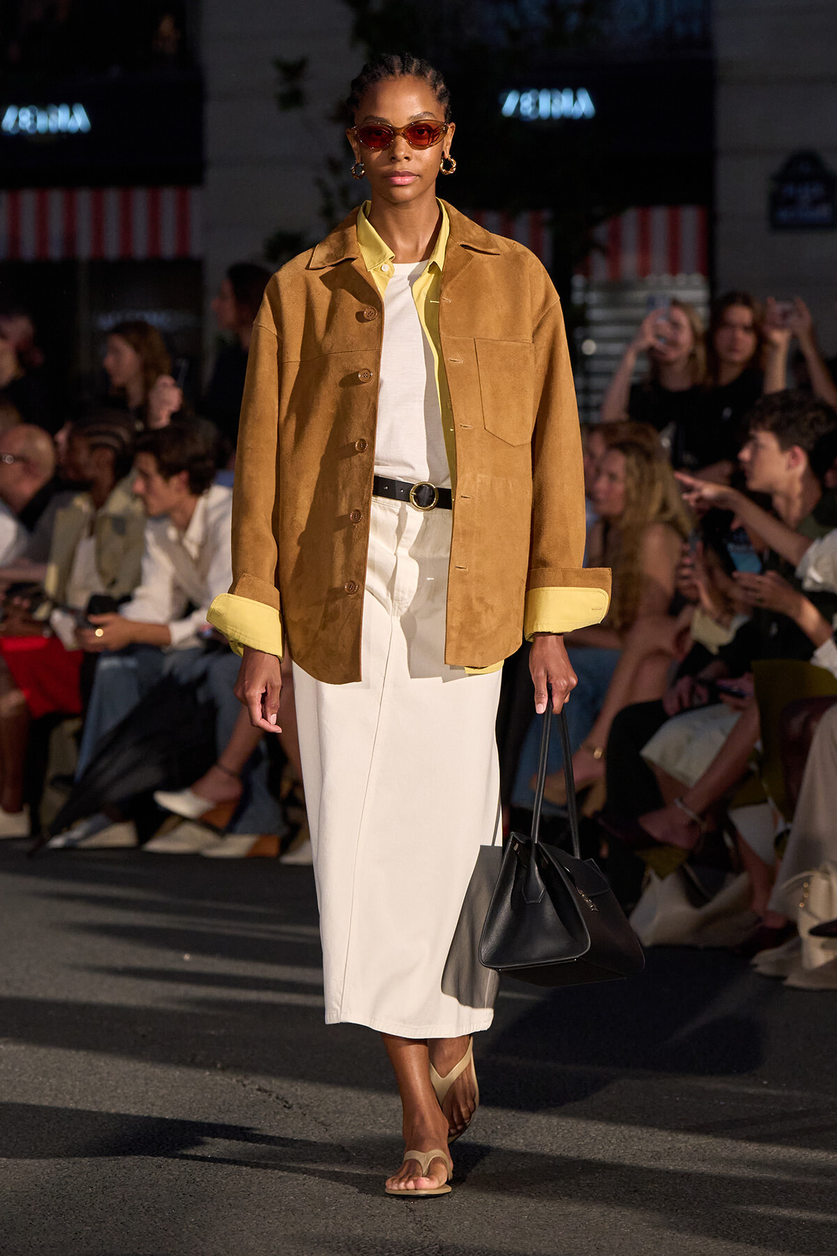 Model walking a runway in a tan suede jacket over a pale yellow shirt, white midi dress, black belt, beige sandals, and red sunglasses while carrying a black handbag.