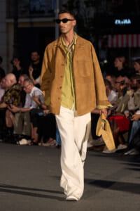 Male fashion model on a runway wearing a tan suede jacket, olive shirt with scarf, white wide-leg pants, sunglasses, and a tan bag in hand.