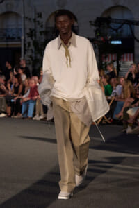 Male model walks a runway in an oversized ivory top with a striped collar, layered sheer jacket pieces, and beige wide-leg pants; audience in the background.