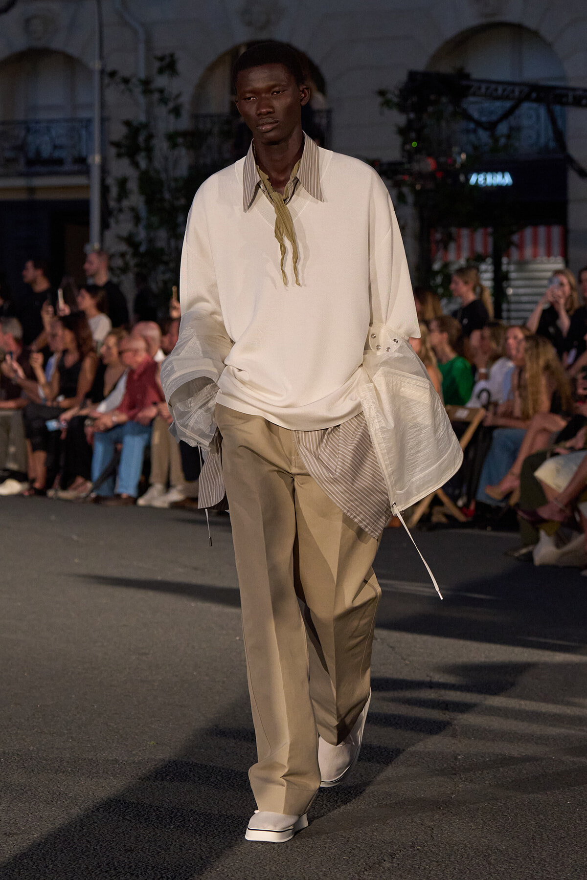 Male model walks a runway in an oversized ivory top with a striped collar, layered sheer jacket pieces, and beige wide-leg pants; audience in the background.