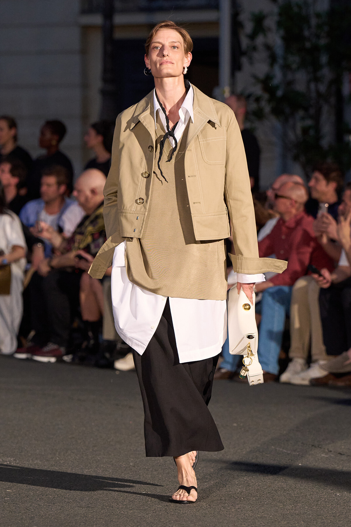 Model walking a runway in a beige cropped jacket over a white shirt with long tails and black wide-leg pants, holding a small white bag in one hand.