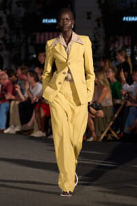 Female model walks a runway in a pale yellow pantsuit with oversized beige collar lapels and matching wide-leg pants, audience in background.