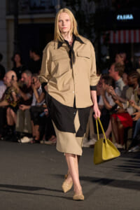 Model walking the runway in a beige and black color-block outfit, carrying a yellow handbag.