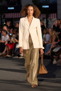 Model walking a runway in a cream blazer, matching wide-leg beige leather pants, and a brown handbag.
