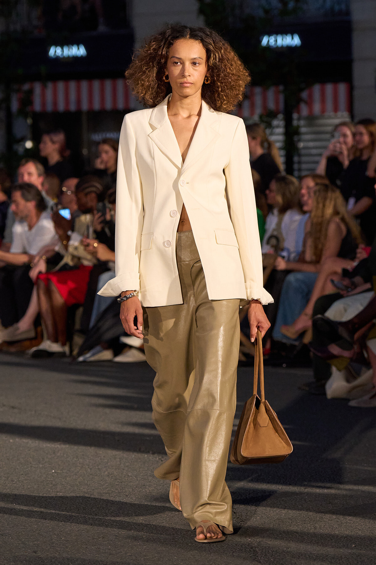 Model walking a runway in a cream blazer, matching wide-leg beige leather pants, and a brown handbag.