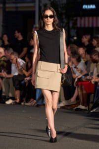 Model walking a runway in a black sleeveless top, beige utility mini skirt with large pockets, black heels, sunglasses, and a black shoulder bag.