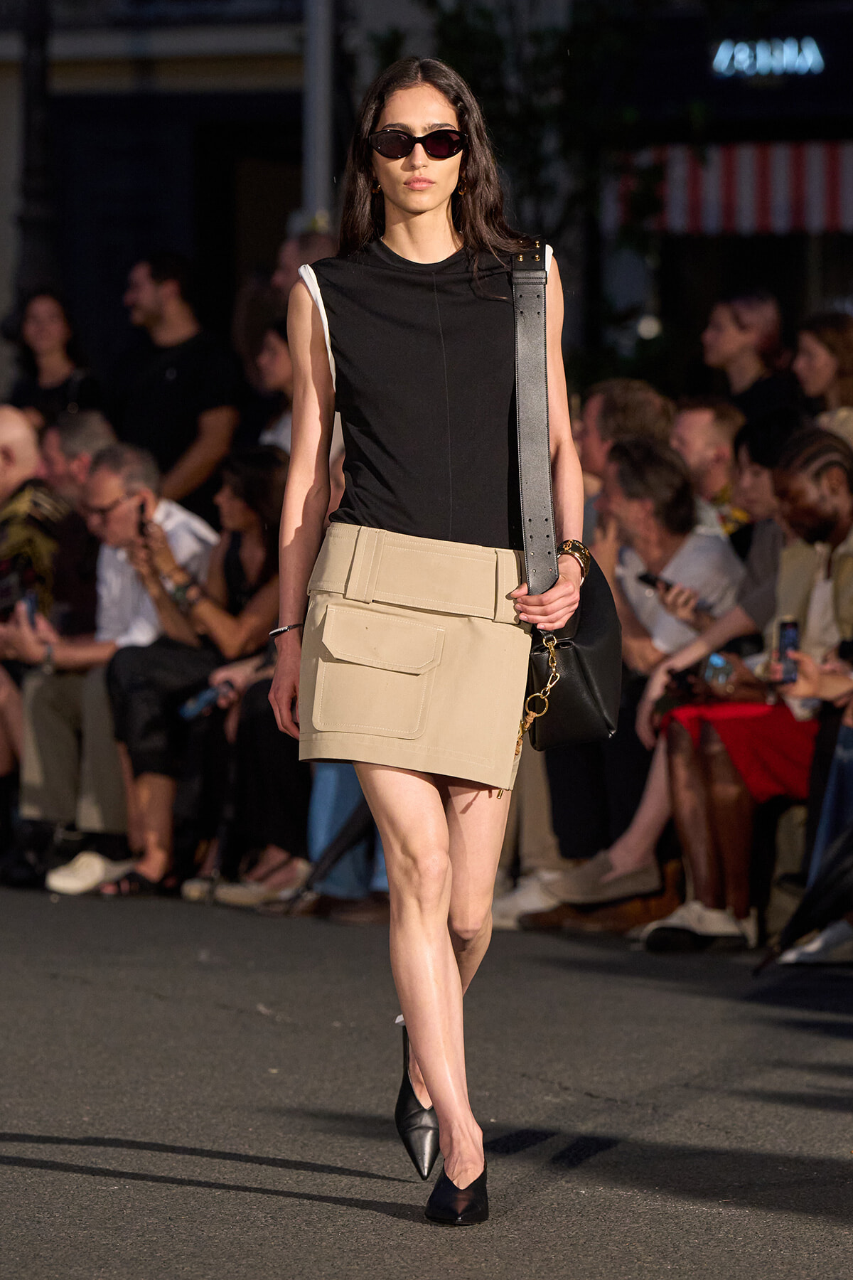 Model walking a runway in a black sleeveless top, beige utility mini skirt with large pockets, black heels, sunglasses, and a black shoulder bag.
