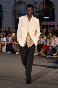 Male model on a runway wearing a cream blazer over a beige shirt and black trousers, spectators seated along the sides.