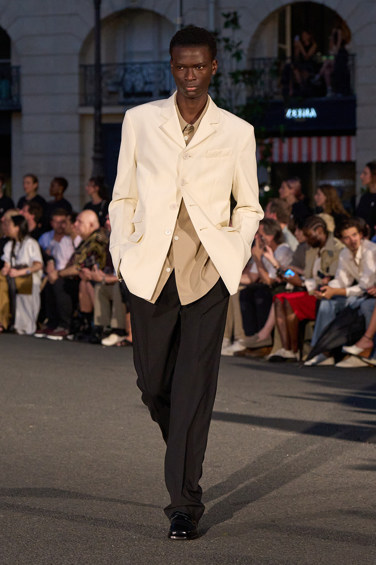 Male model on a runway wearing a cream blazer over a beige shirt and black trousers, spectators seated along the sides.