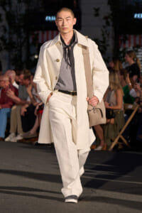 Male model walking a fashion runway in a cream trench and matching wide-leg pants, gray top, black scarf, and a beige crossbody bag, with an audience in the background.