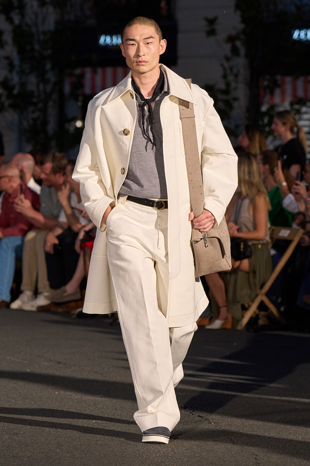 Male model walking a fashion runway in a cream trench and matching wide-leg pants, gray top, black scarf, and a beige crossbody bag, with an audience in the background.